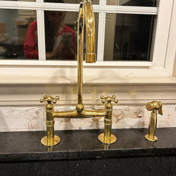 Gold kitchen faucet set on a black countertop with a window in the background.